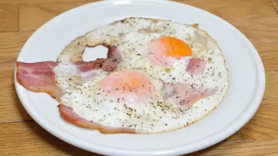 Two fried eggs with bacon and black pepper on a white plate on a wooden table