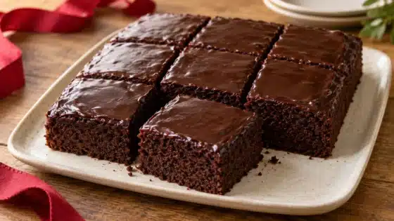 Nine glazed square portions of chocolate cake on a pale platter with a red ribbon on the table